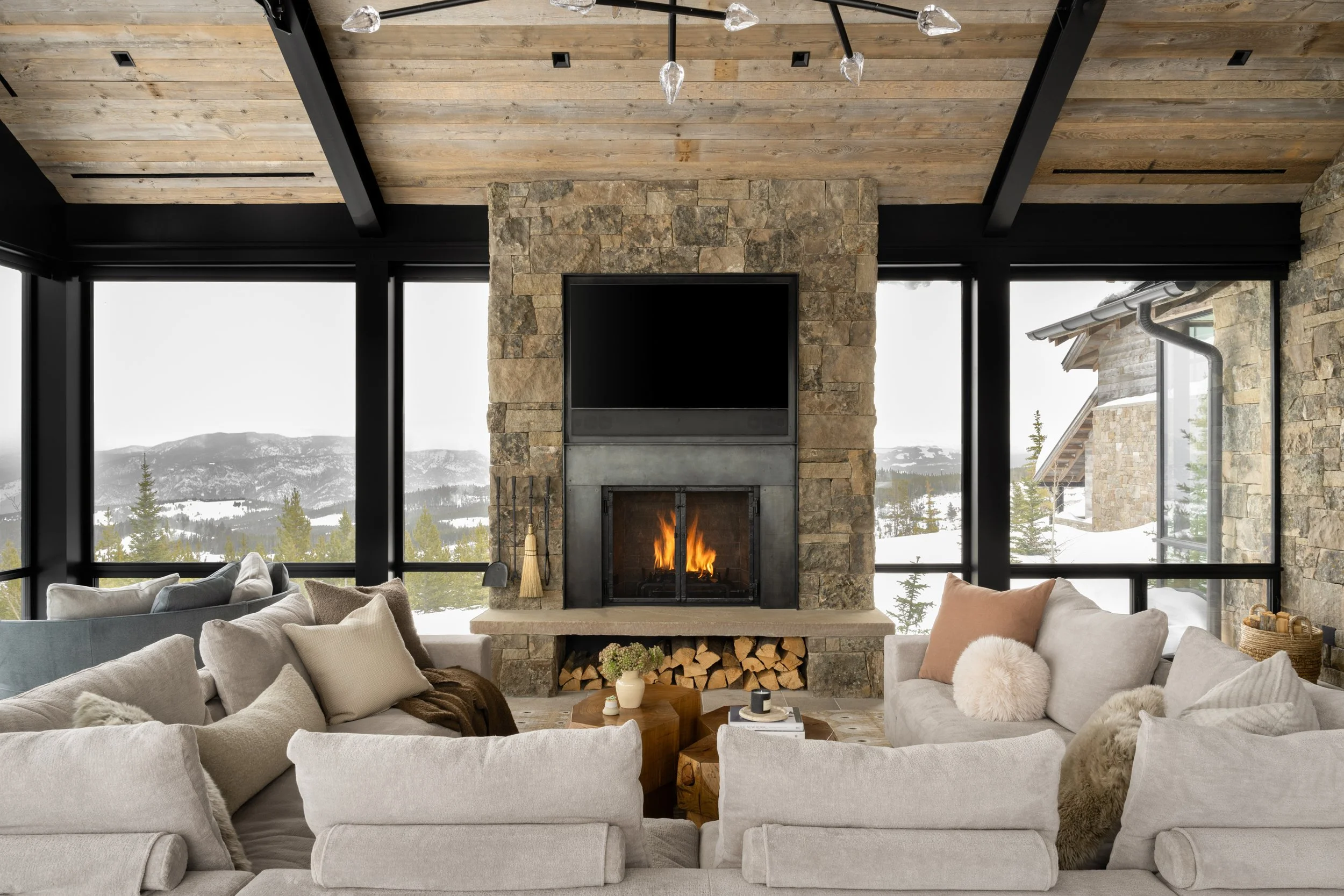 Discover the ultimate mountain retreat. A stunning rustic modern living room features a majestic stone fireplace, cozy neutral seating, and dramatic floor-to-ceiling windows with panoramic snowy mountain views. Perfect for luxury interior design and architecture inspiration.